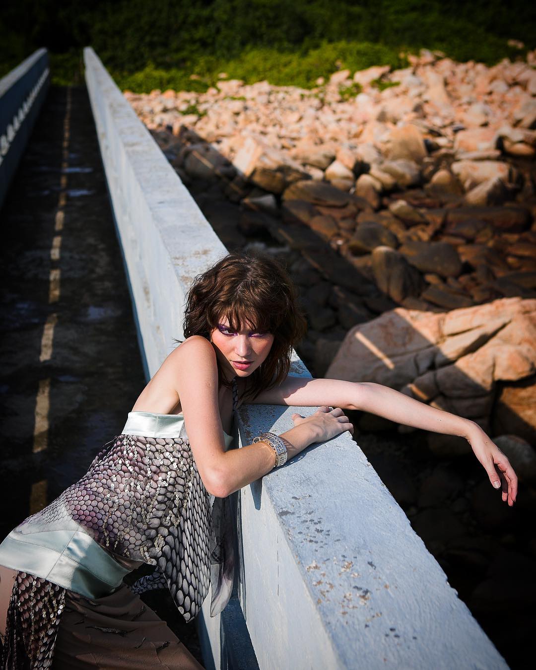 Fashion model reclining across a bridge above a rocky shoreline